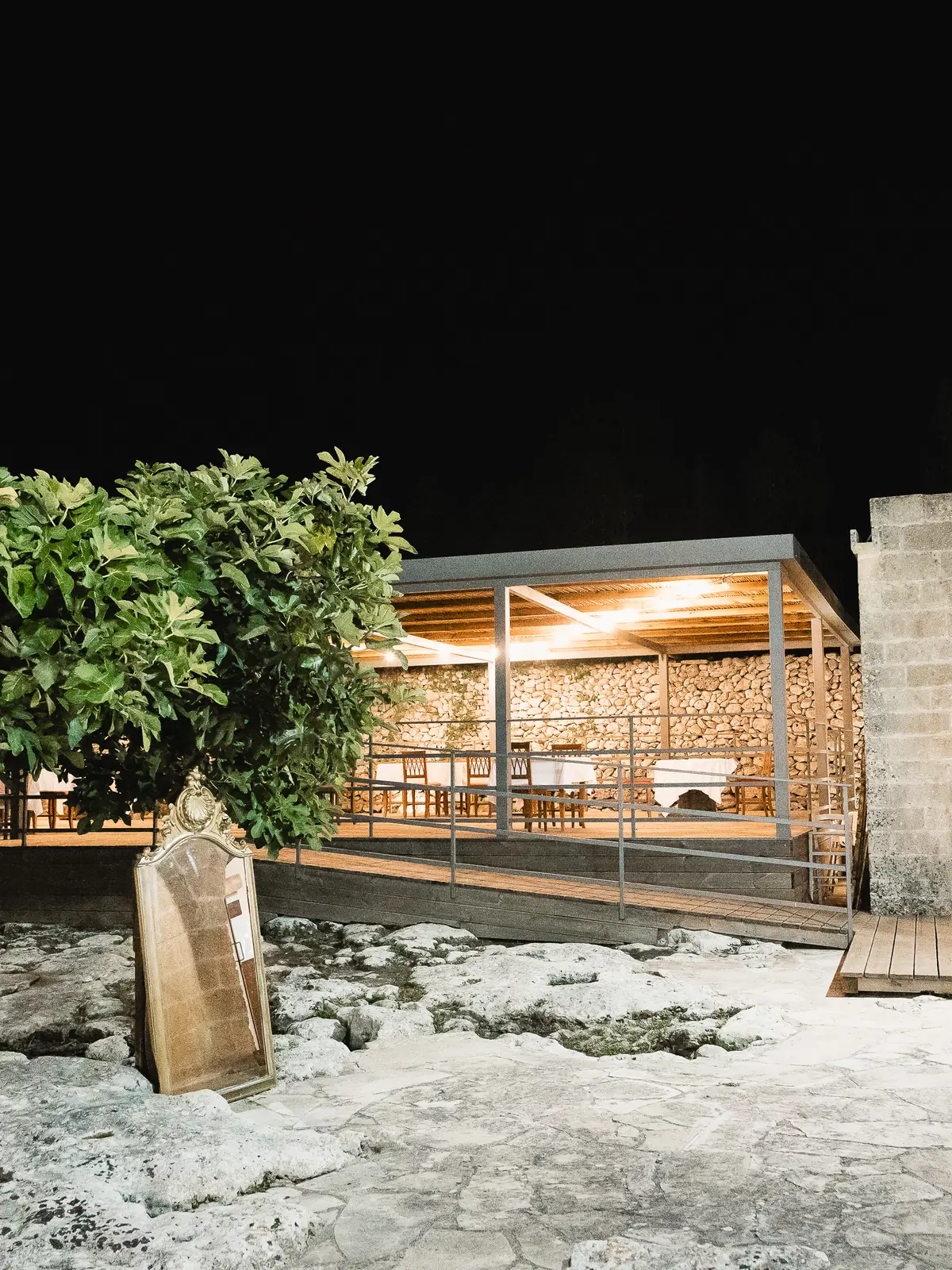 La loggia della masseria illuminata la sera, con le tavolate pronte, oltre il fico